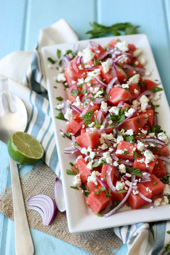 watermelon mint feta salad on a white plate, next to salad servers, lime wedge and mint sprig