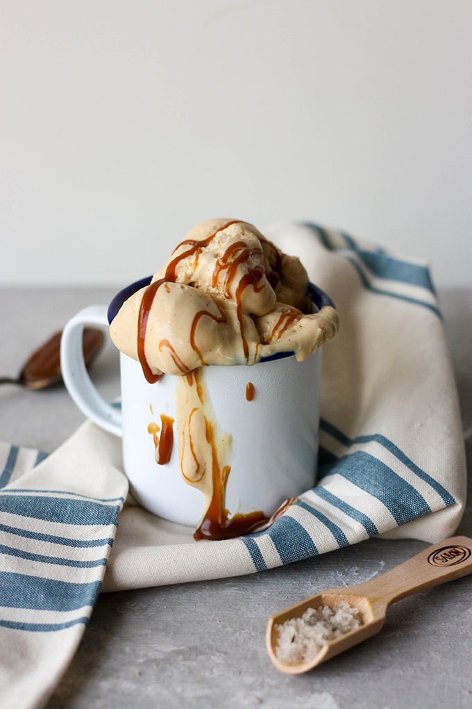 no churn ice cream in white and blue enamel mug with ice cream melted and salted caramel dripping down sides of mug. mug placed on a white and blue napkin next to wooden spoon full of vanilla salt