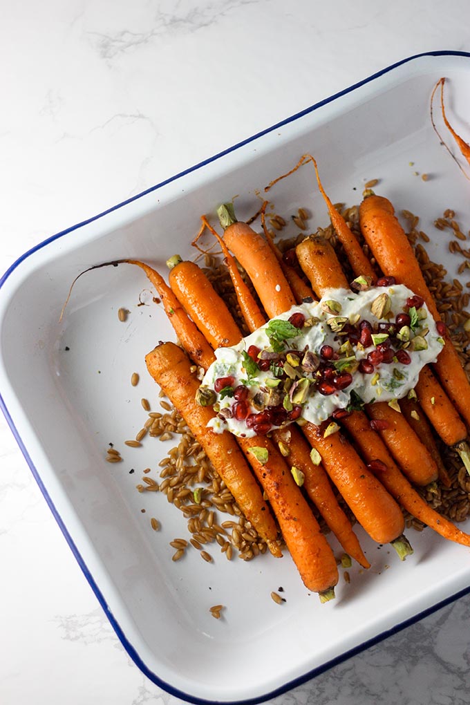 roasted harrisa carrots on farro in a white and blue enamel baking dish, topped with mint yogurt, pistachio and pomegranate seeds. 