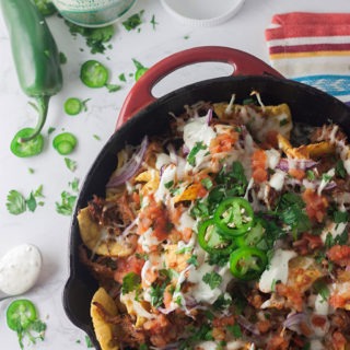 red skillet full of pulled pork nachos, jar of ranch dressing and jalapenos on white board