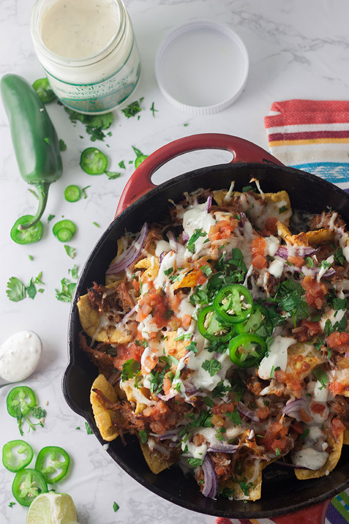 red skillet full of pulled pork nachos, jar of ranch dressing and jalapenos on white board