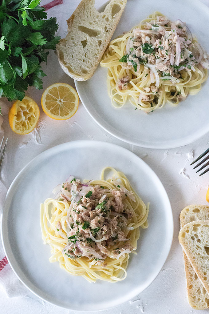 overhead tuna lemon pasta on white plate, white background, bread in bottom right corner, parsley and lemon in top left