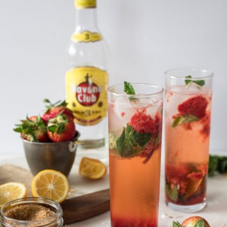 two tall glasses of strawberry mojitos, havana club bottle in background, lemonand strawberries on marble board