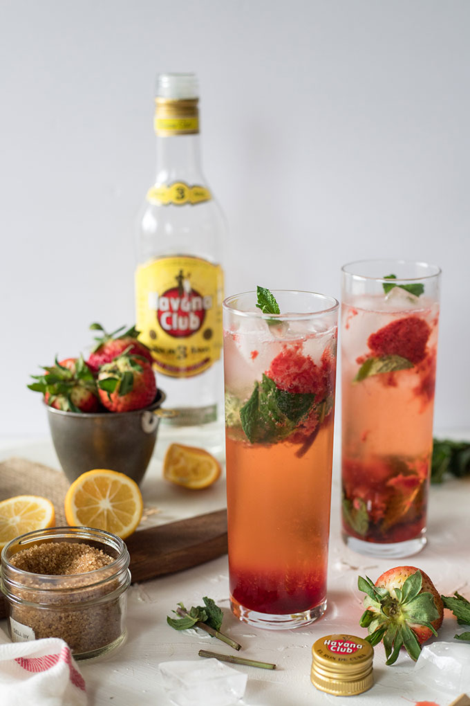 two tall glasses of strawberry mojitos, havana club bottle in background, lemonand strawberries on marble board