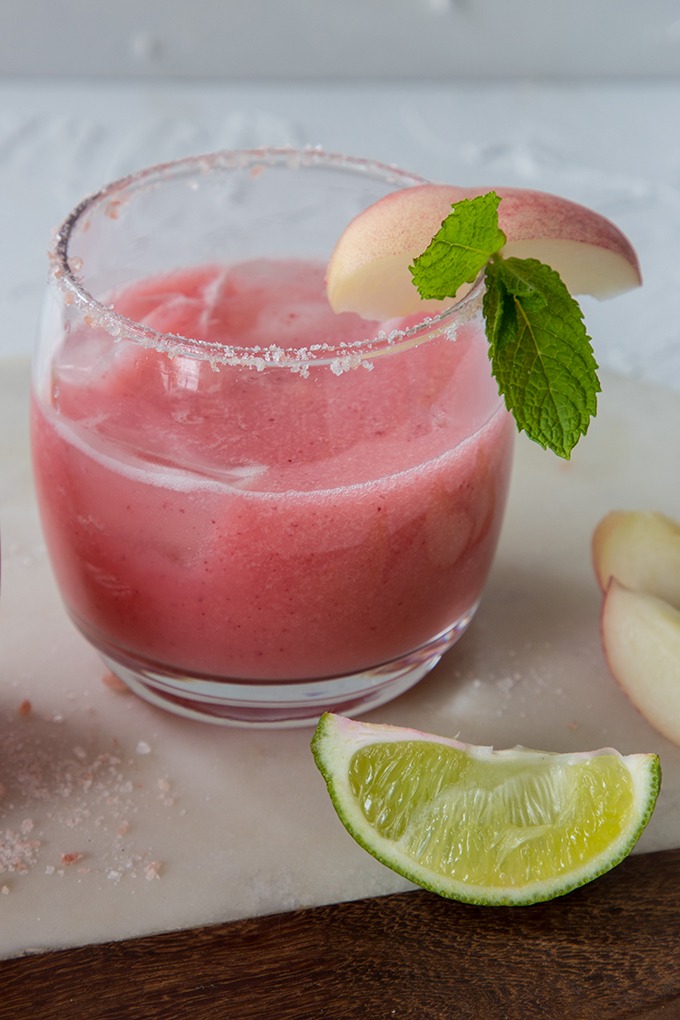 peach margarita in glass on a white marble board, lime wedges and peach wedges littered around glass