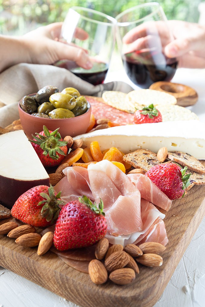 meat and cheese board with two people holding wine glasses doing cheers in the background