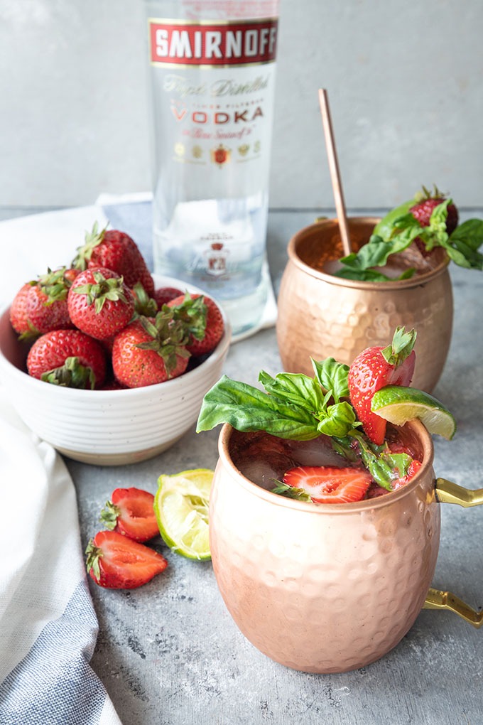 two strawberry basil mule mugs in front of bowl of strawberries and Smirnoff vodka bottle