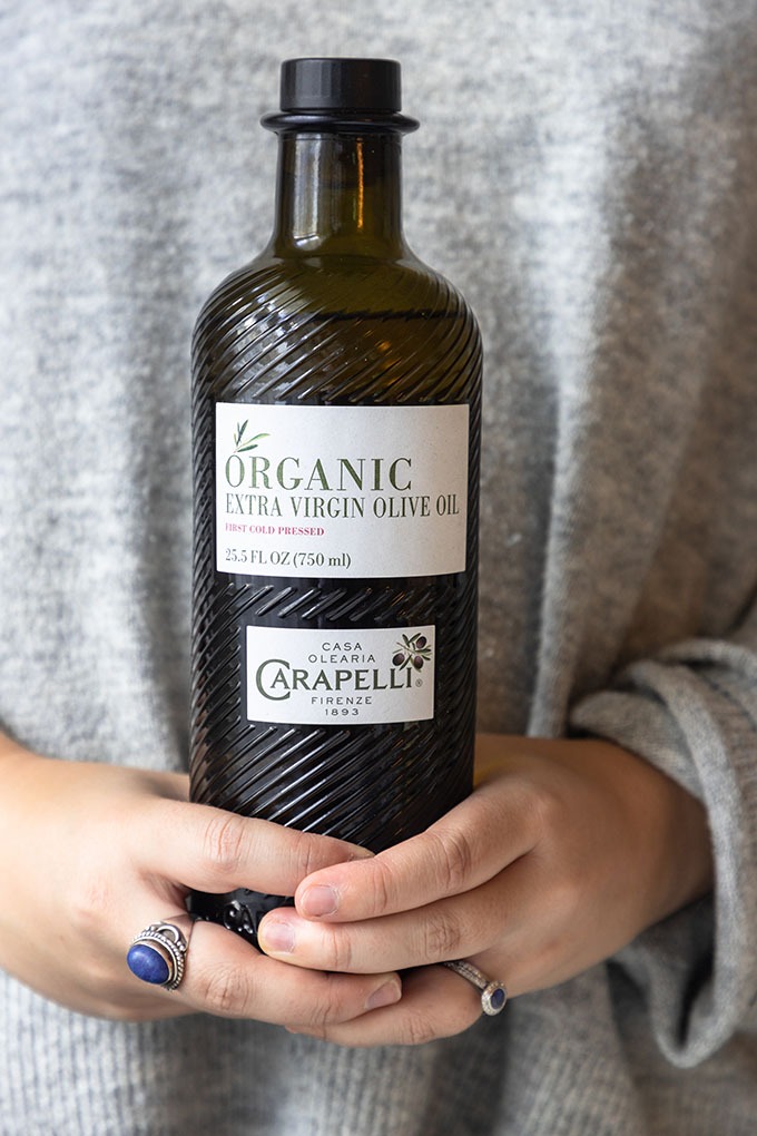 person in grey sweater holding bottle of carapelli olive oil