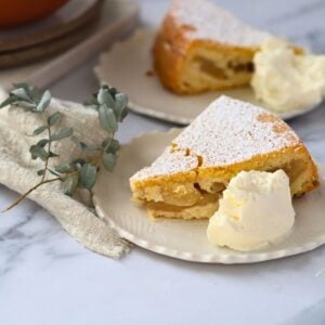 piece of apple cake on a small plate with a dollop of whipped cream to the side of the cake. A second plate of cake is behind the front plate with a small eucalyptus sprig.