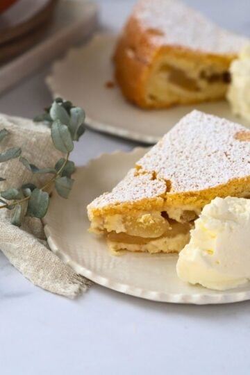 piece of apple cake on a small plate with a dollop of whipped cream to the side of the cake. A second plate of cake is behind the front plate with a small eucalyptus sprig.