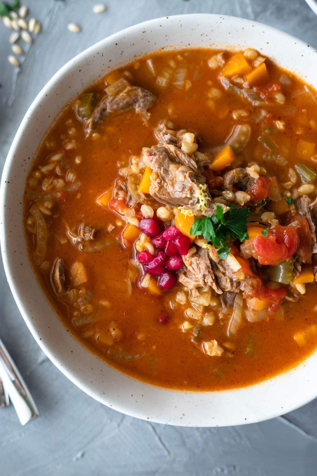Lamb shank soup in a bowl topped with pomegranate, lemon and parsley. Left of the bowl is cutlery and there are small pieces of barley around the bowl.