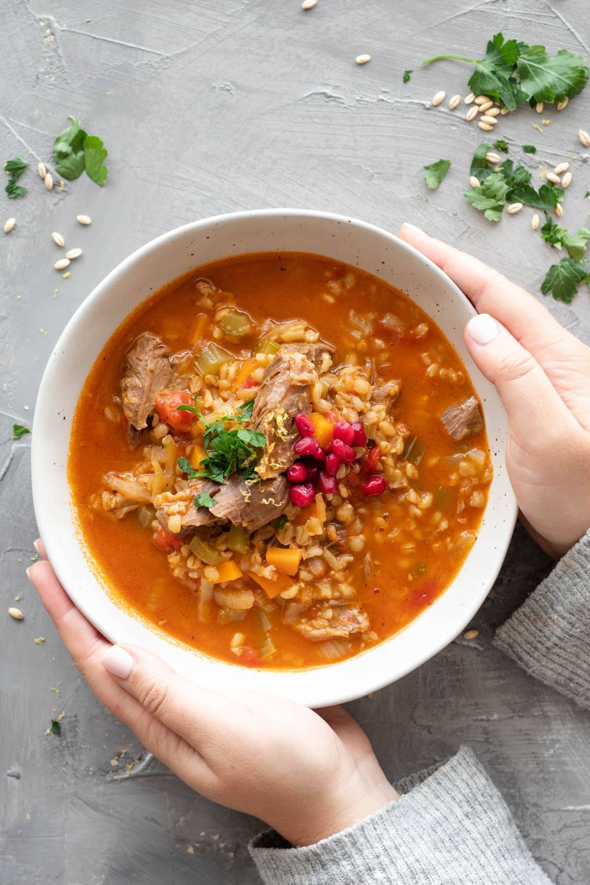 Two arms wearing a sweater reaching out to hold a bowl of lamb shank soup. There is loose parsley and barley on the backdrop.