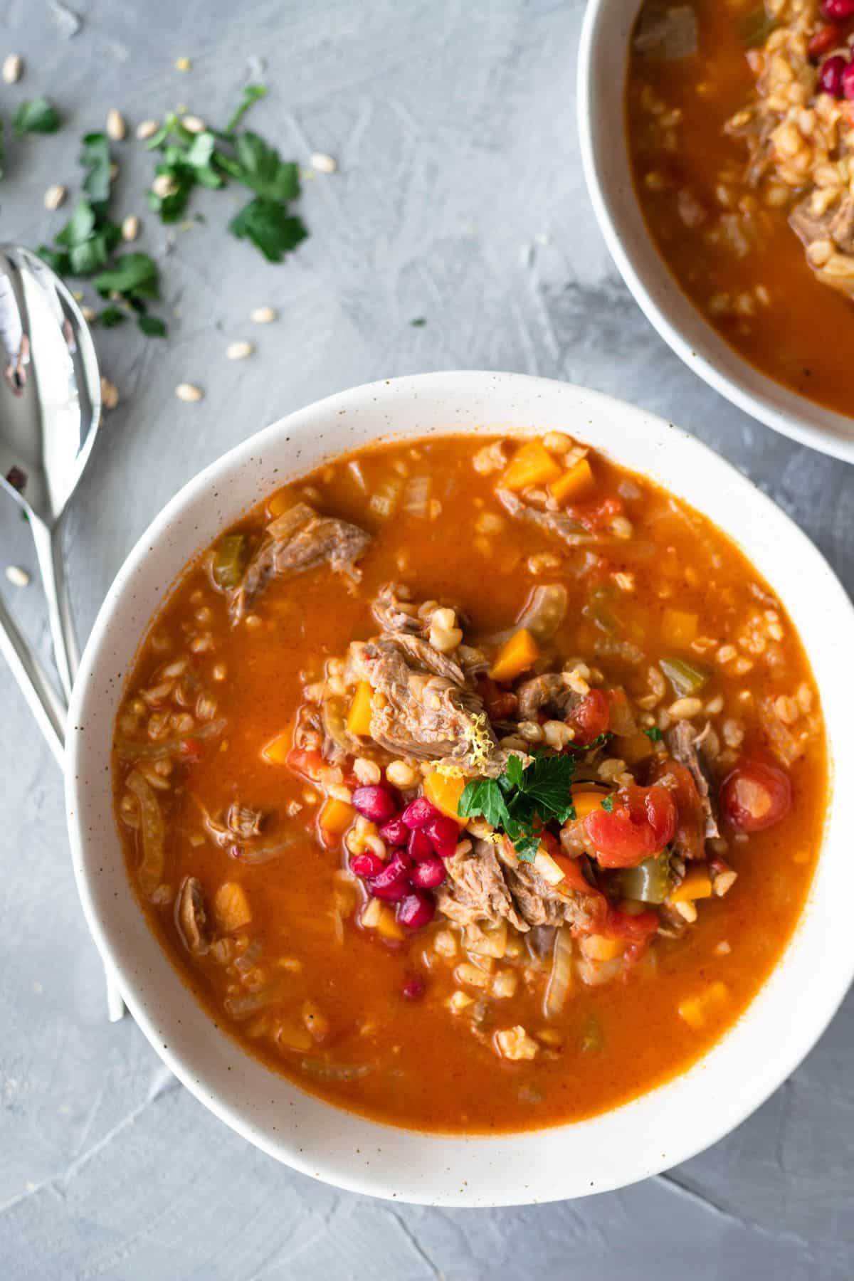 lamb shank soup in white bowl. There is also cutlery next to the bowl and loose barley and parsley on the top of the background.