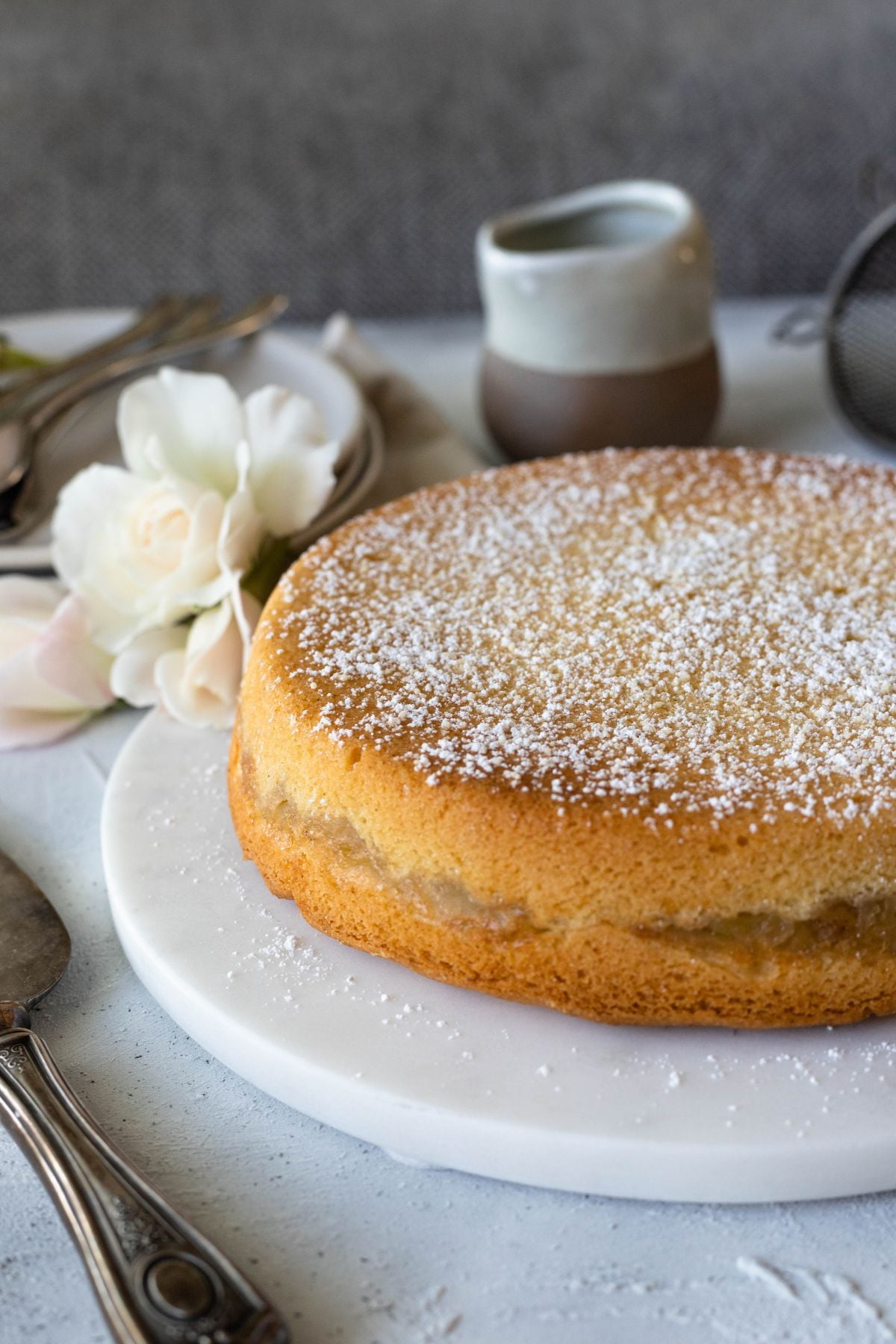 whole cake on a circular board. A small pitcher sits behind the cake with some white roses.