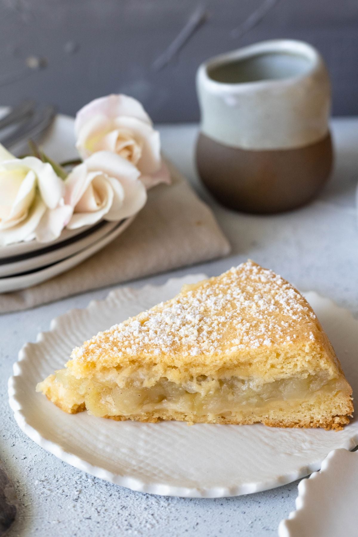 a slice of cake on a rustic plate. There is a small pitcher behind the cake with some white roses.