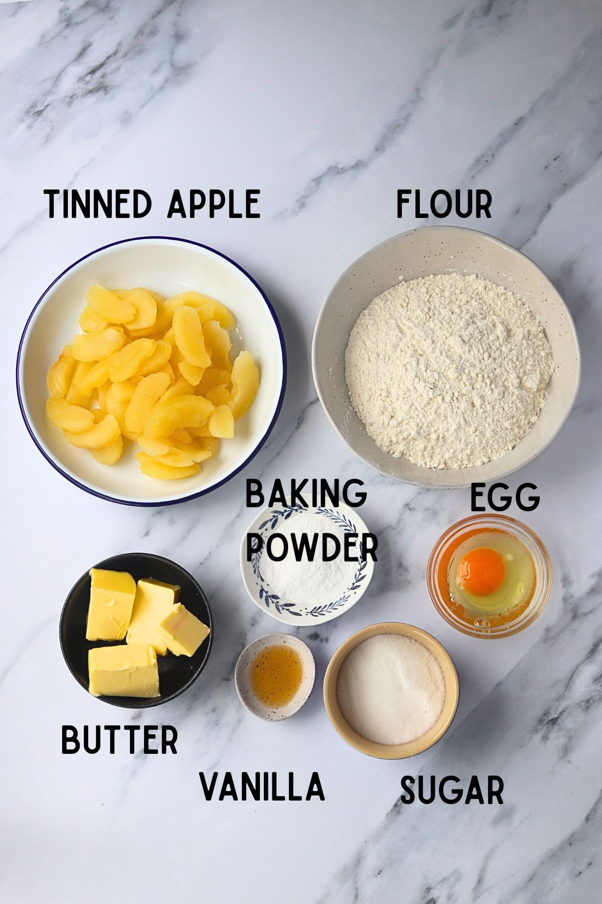 ingredients laid out on a marble board. All ingredients in bowls. Top to bottom left to right - apple, flour, butter, baking powder, egg, vanilla, sugar.