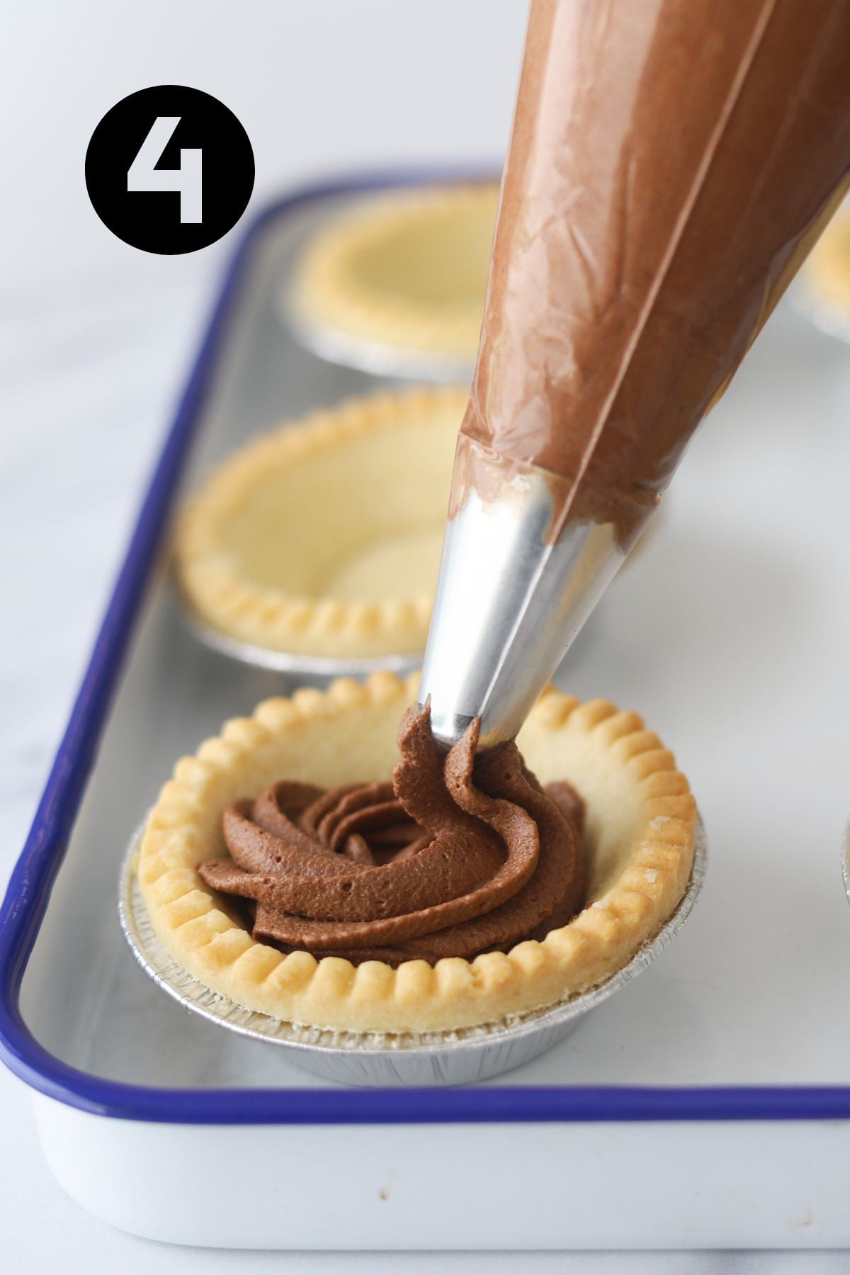 chocolate mousse being piped into a tart shell. tart shells are sitting in an enamel tray.