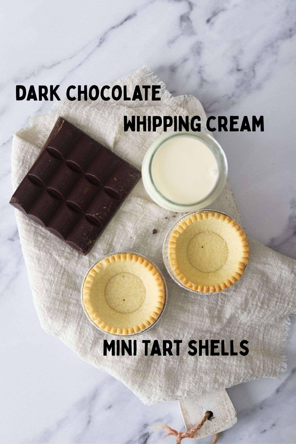 dark chocolate, cream in a glass jar, two empty tart shells on a chopping board placed on a linen napkin.