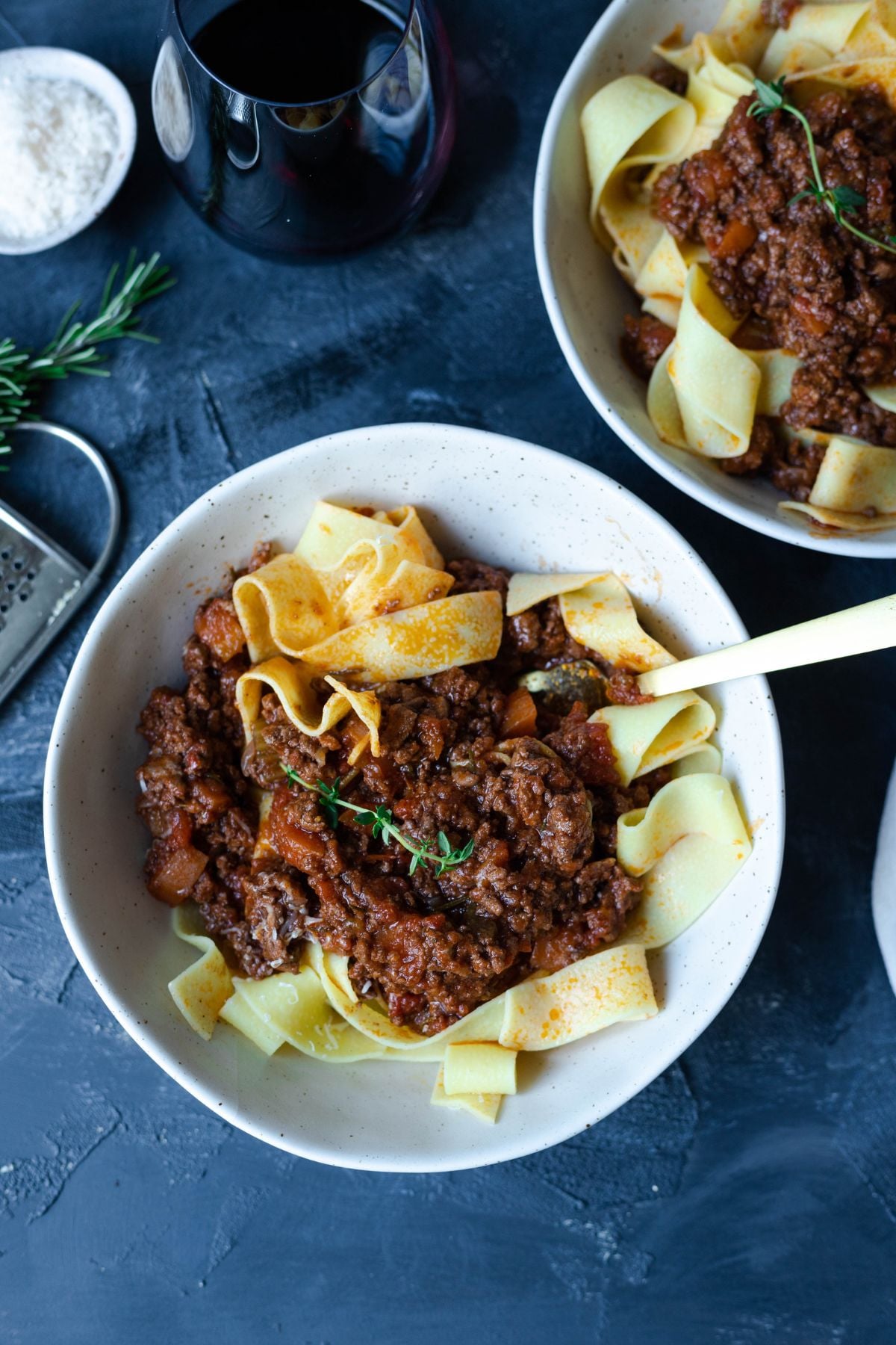 lamb ragu and papardelle in a small bowl on a board. there is a fork in the bowl, a glass of wine at the top of the image and a small dish of salt next to the wine.