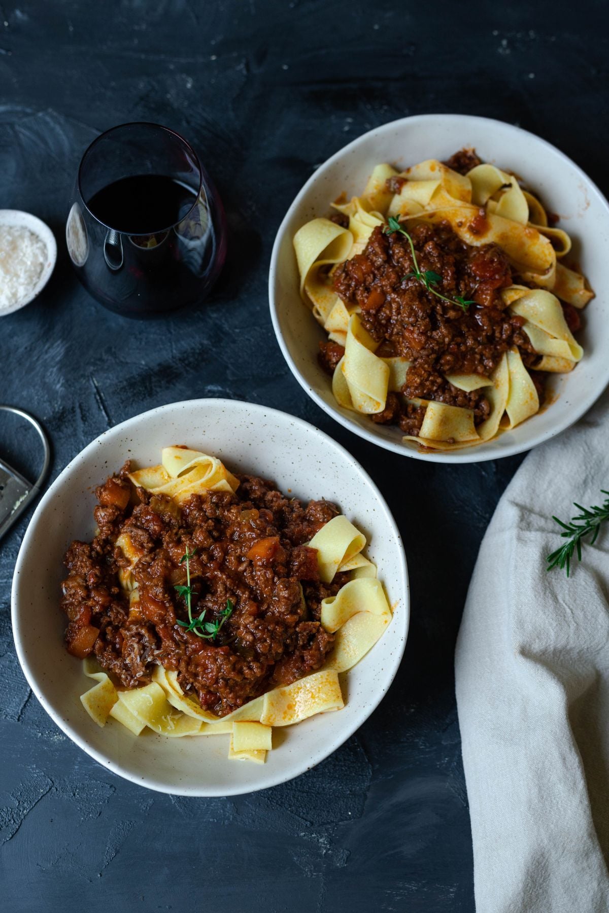 two bowls of lamb ragu with pasta on a board. A glass of wine is next to one of the bowls of pasta and there is napkin on the board too.