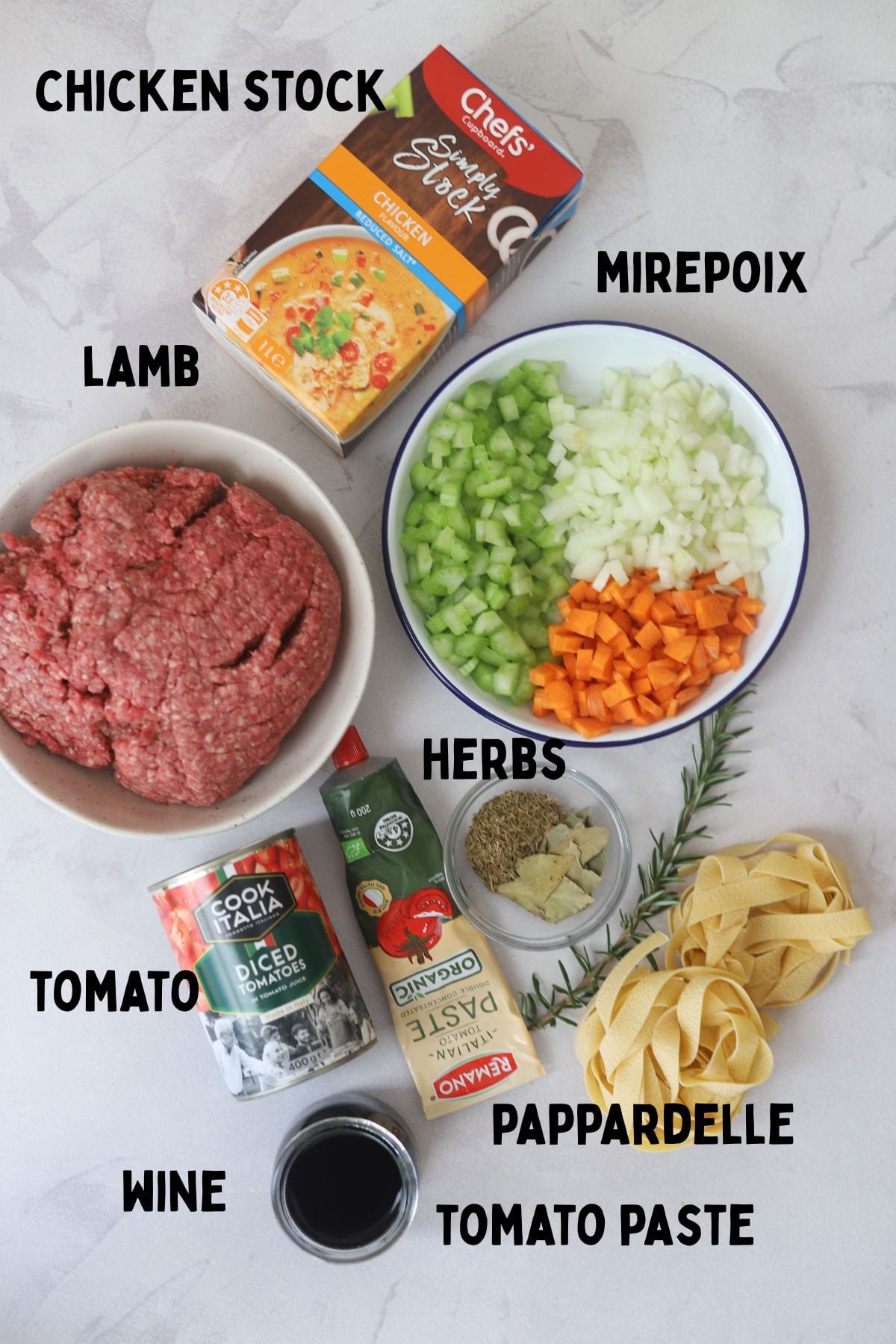 lamb ragu ingredients laid out on board. From top left to right. Lamb mince in bowl, carton of chicken stock, celery, carrot and onion in a bowl. Tin of tomatoes, tomato pasta, herbs, pappardelle and red wine.