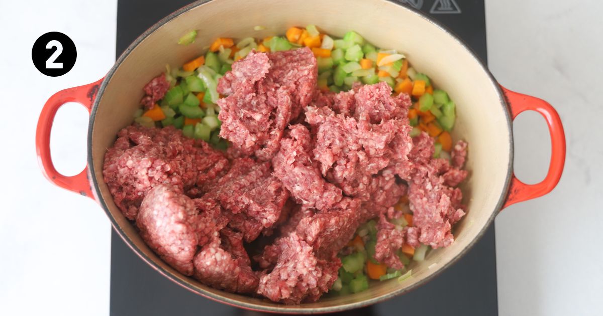 raw lamb mince on top of onion, carrot and celery in dutch oven on a portable stove top.