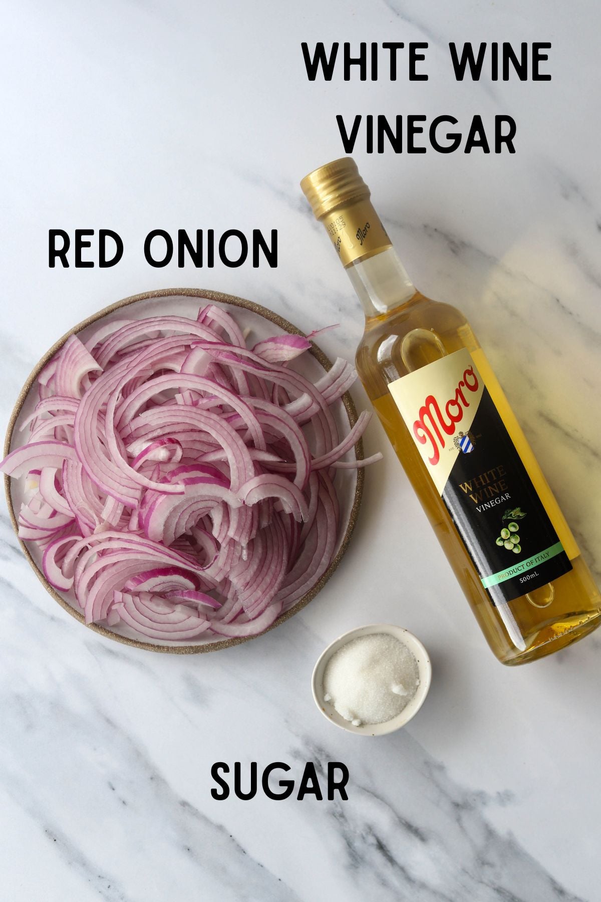 3 ingredients for pickled onions on a marble board. Red onion on a small plate, a bottle of white wine vinegar and a small bowl of sugar pictured.