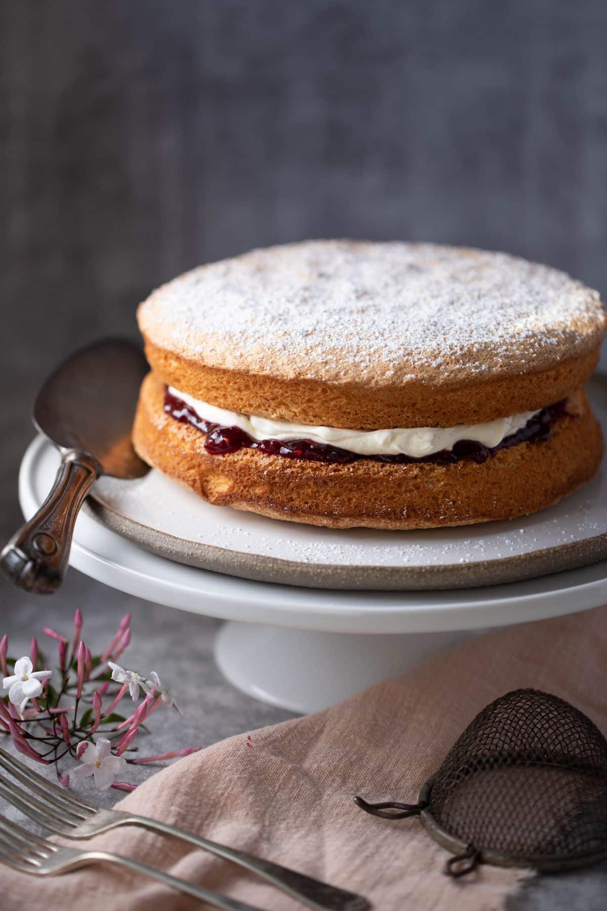 sponge cake on a cake stand. Cake is filled with jam and cream and dusted with icing sugar. There is some jasmine below the cake stand and a linen cloth.