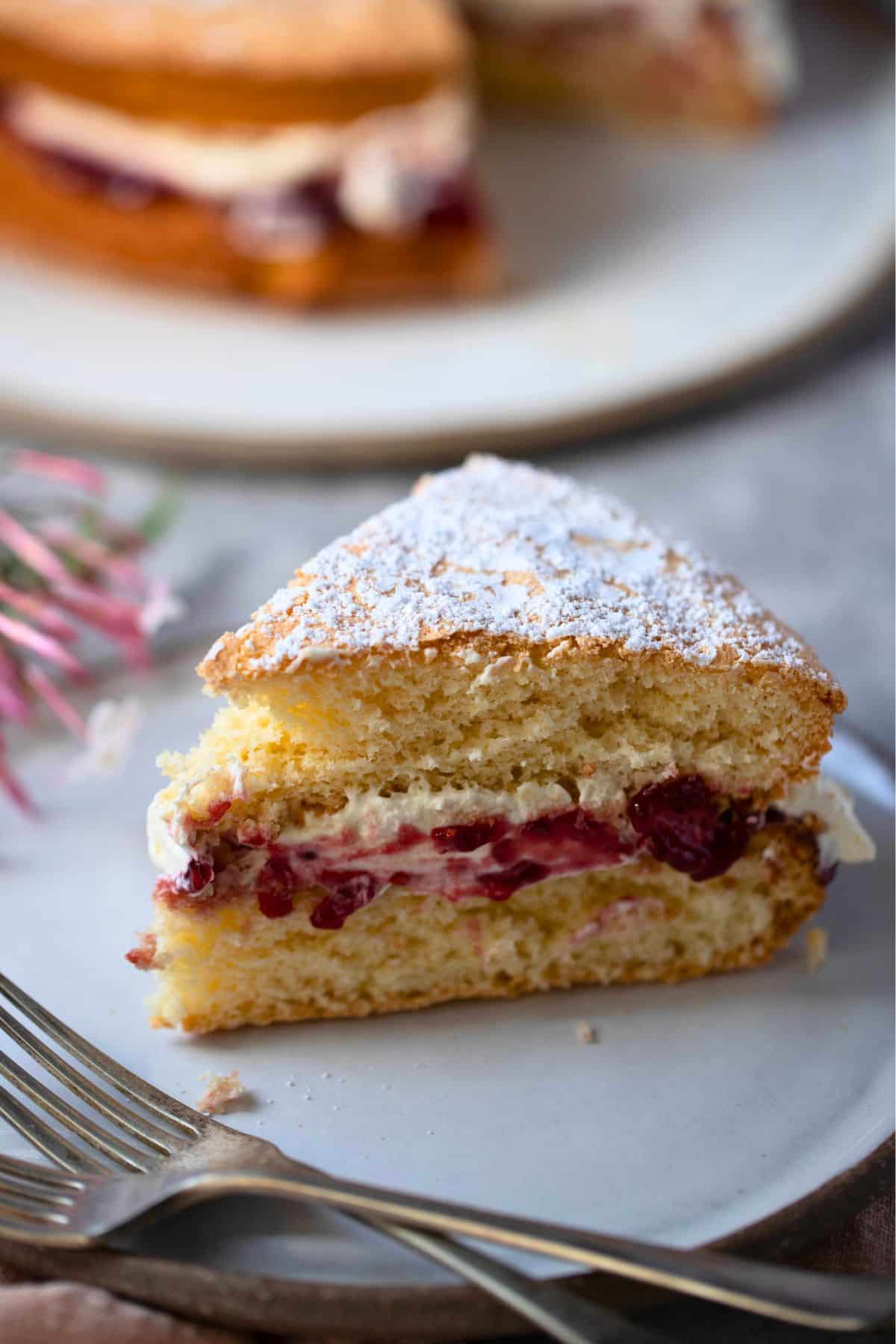 slice of sponge cake on a small plate with old fashioned cutlery and jasmine flours on the side of the plate.