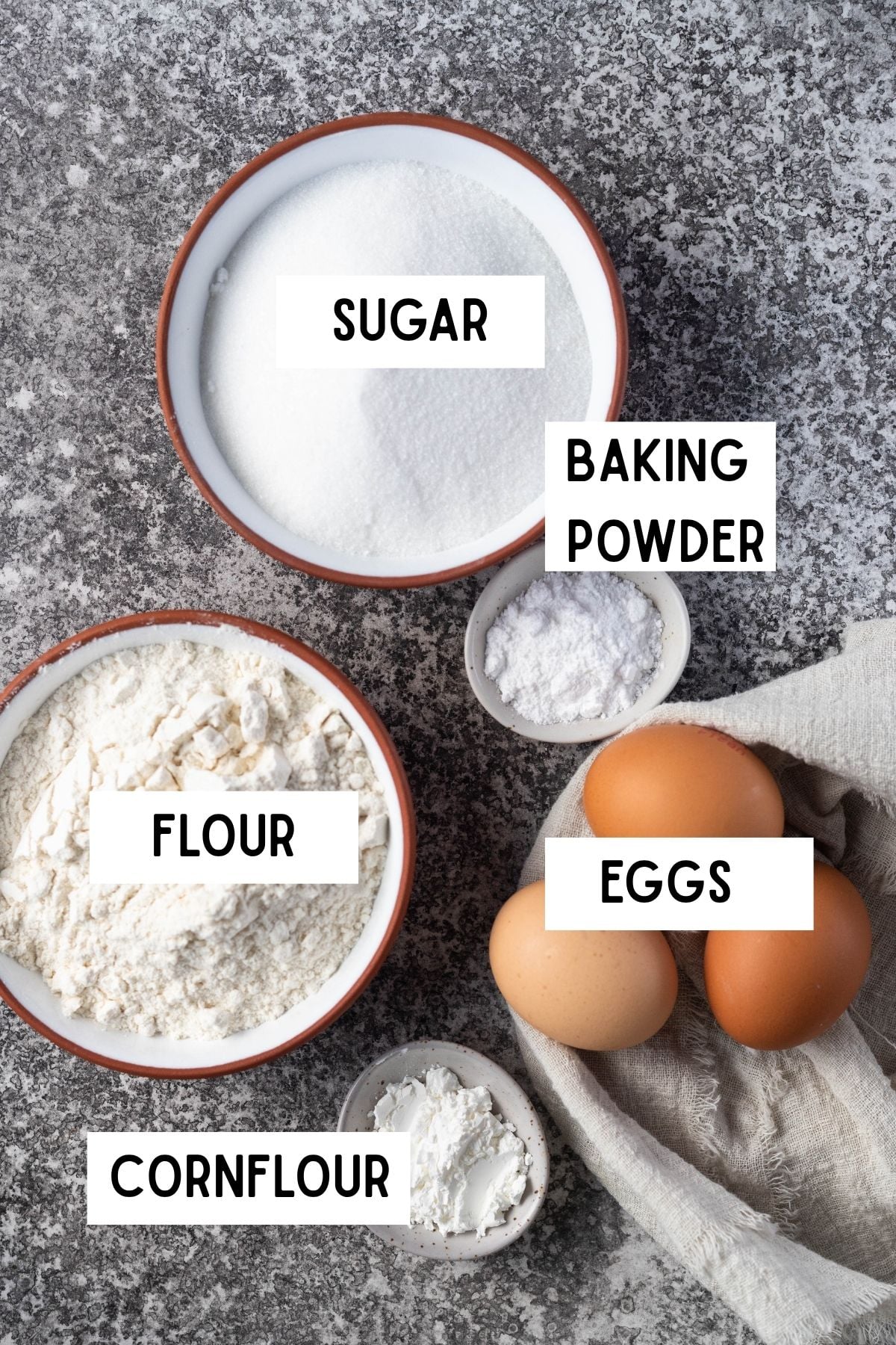 sponge cake ingredients laid on photography board. From top to bottom, left to right - sugar, flour, baking powder, cornflour and eggs.