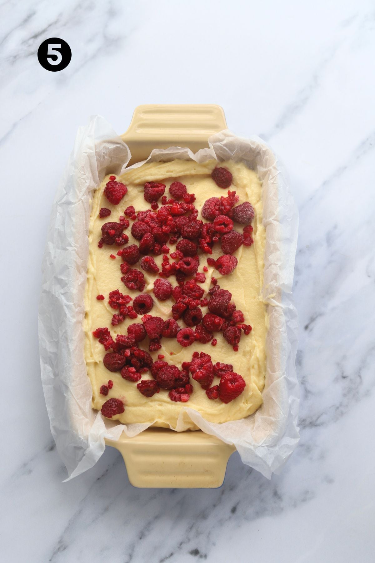 Raspberries scattered onto batter which is in a long, rectangular baking dish lined with parchment paper.