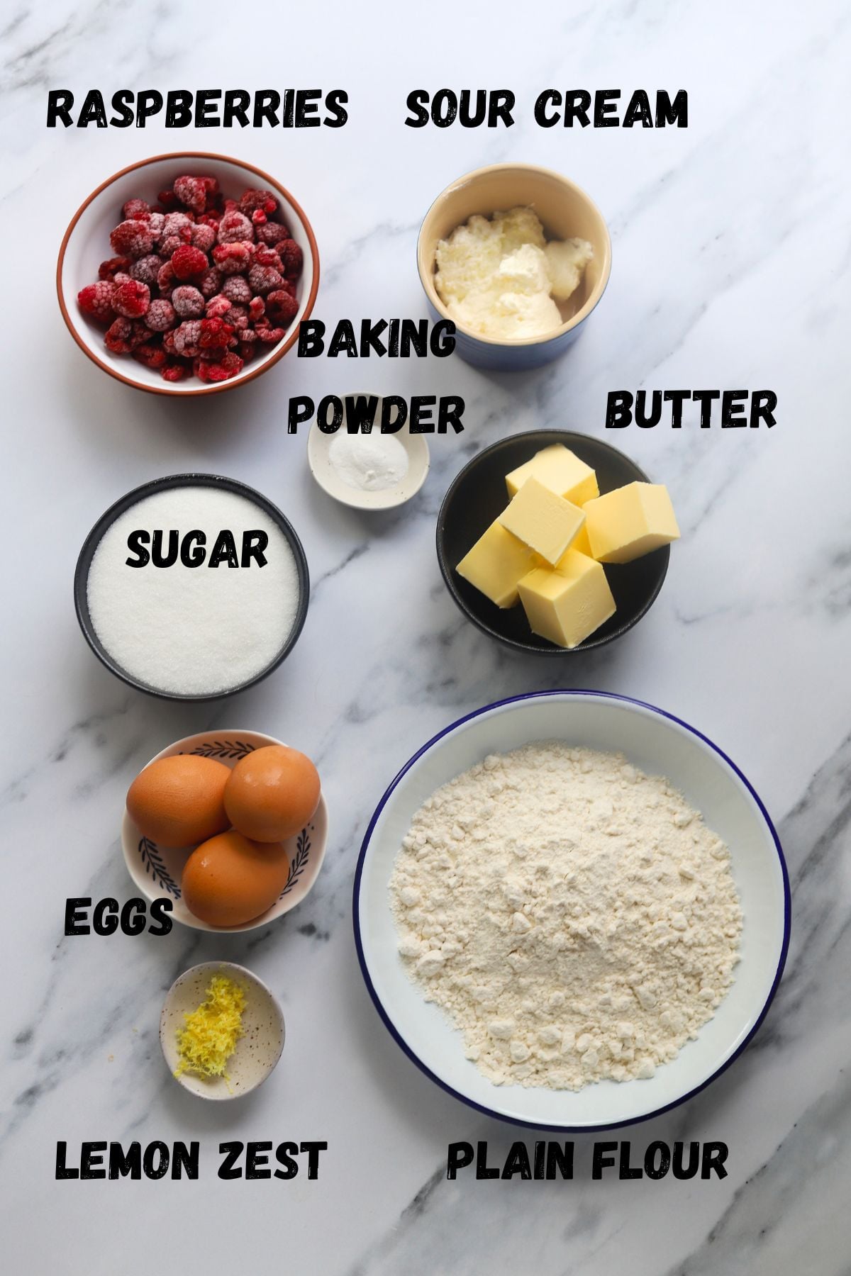 lemon cake key ingredients on a board. From top to bottom, left to right - raspberries, sour cream, sugar, baking powder, butter, eggs, lemon zest and flour. All ingredients in small bowls.