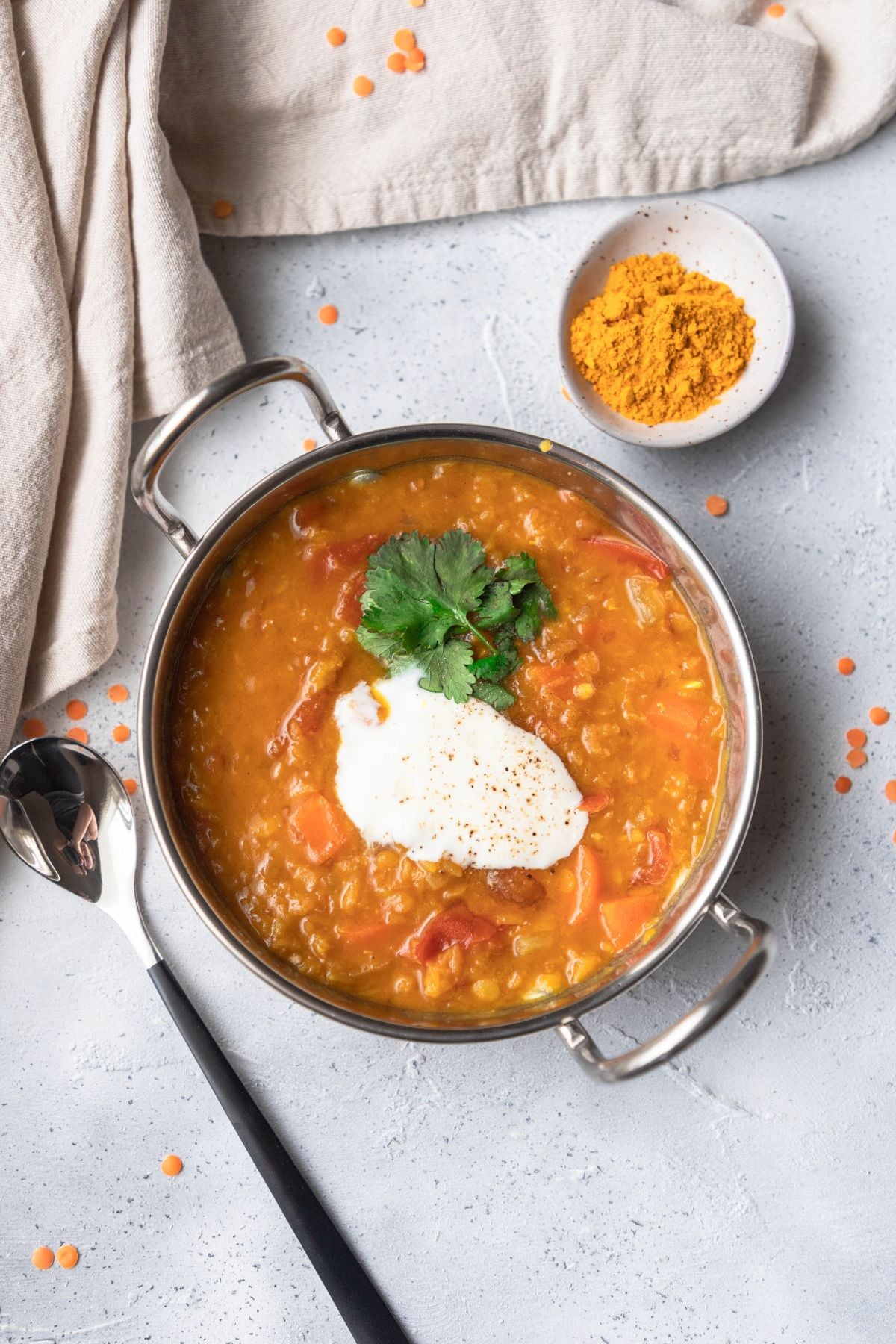 A small bowl of lentil dal on a board. A spoon is to the left of the bowl and a small bowl of red lentils to the right. There are scattered lentils around the bowls.